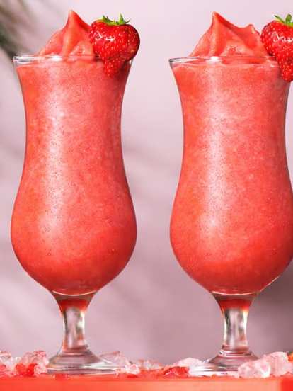 Malibu Strawberry Colada cocktails in hurricane glasses with fresh strawberry garnish, placed on a red tray with ice cubes and sliced strawberries beside a red blender against a soft pink background with green leaves.