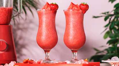 Malibu Strawberry Colada cocktails in hurricane glasses with fresh strawberry garnish, placed on a red tray with ice cubes and sliced strawberries beside a red blender against a soft pink background with green leaves.