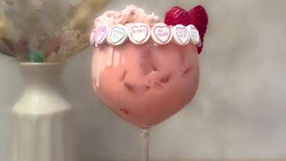 Malibu Strawberry Colada Float in a large stemmed glass with creamy pink drink, ice, candy hearts around rim, and fresh strawberries garnish, placed on wooden surface with neutral background.