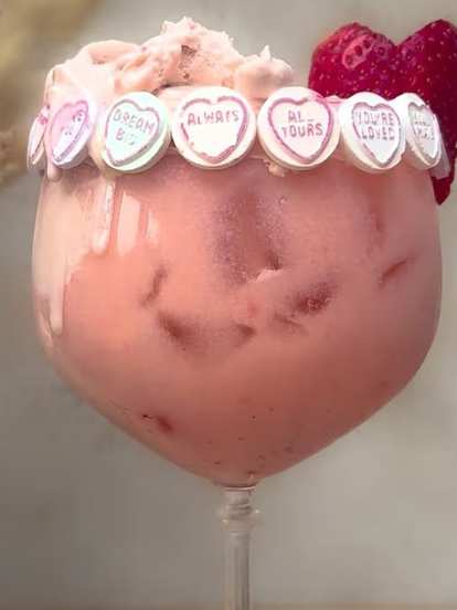 Malibu Strawberry Colada Float in a large stemmed glass with creamy pink drink, ice, candy hearts around rim, and fresh strawberries garnish, placed on wooden surface with neutral background.