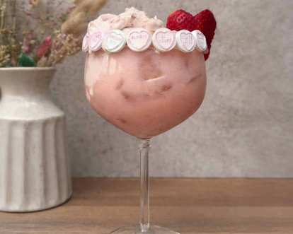 Malibu Strawberry Colada Float in a large stemmed glass with creamy pink drink, ice, candy hearts around rim, and fresh strawberries garnish, placed on wooden surface with neutral background.