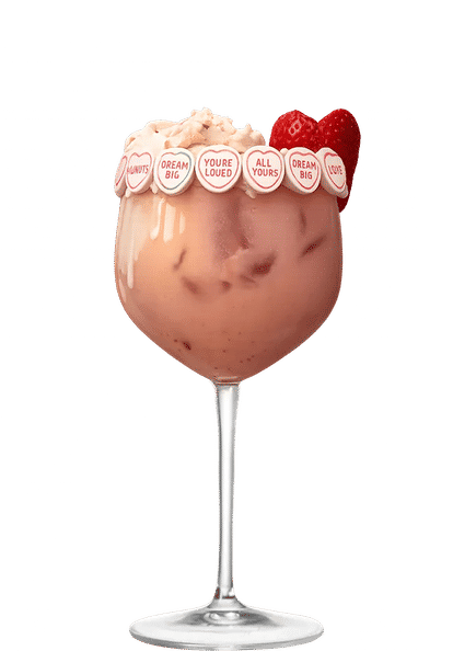 Malibu Strawberry Colada Float cocktail in a large stemmed glass with creamy pink drink, ice cubes, whipped topping, candy hearts around the rim, and a strawberry garnish, isolated on a white background.