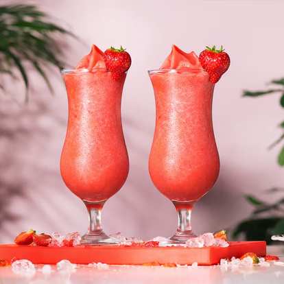 Two Malibu Strawberry Colada cocktails in hurricane glasses with fresh strawberry garnish, standing on a red surface scattered with ice and strawberry pieces against a pink backdrop with green foliage.