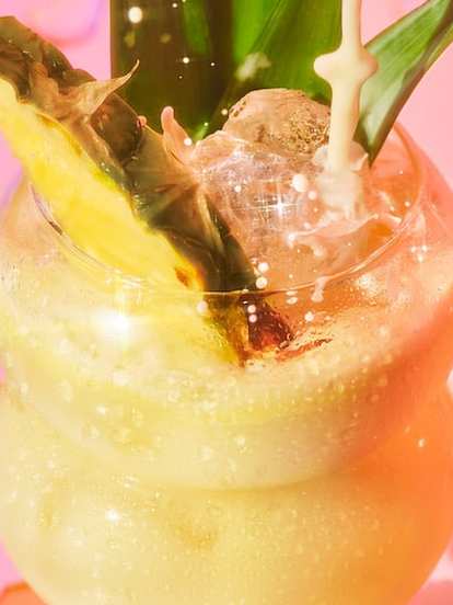 Malibu Dirty Coconut — a creamy tropical cocktail served in a curvy glass with ice, pineapple wedge, and leaves, splashing under warm pink lighting for a rich, beach‑ready look.