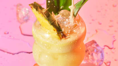 Malibu Dirty Coconut — a creamy tropical cocktail served in a curvy glass with ice, pineapple wedge, and leaves, splashing under warm pink lighting for a rich, beach‑ready look.