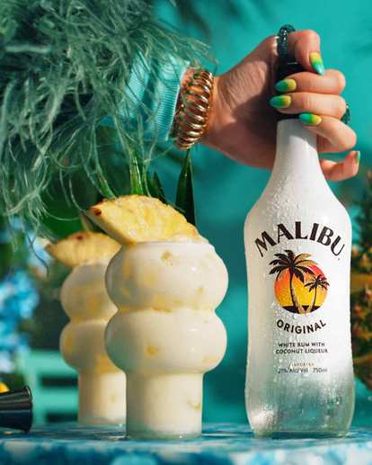 A bottle of Malibu and two pina coladas in a bubbly glass decorated with pineapple garnish